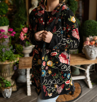 Model standing against a backdrop of potted plants and a bench on a porch deck wears a long black cardigan with colorful embroidered designs and the words, "Hope" and "Love" in white script all over it