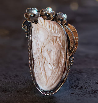 Carved bone necklace pendant features two horse's heads nestled together between feathers and set in an ornate silver backing with floral accents