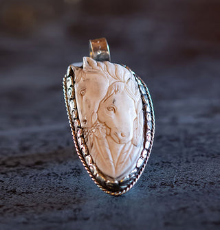 Carved bone necklace pendant features two horse's heads nestled together and framed by feathers inside an ornate silver setting with top-set bail