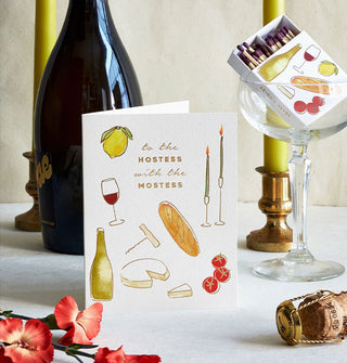To the Hostess With the Mostess greeting card is staged on a tabletop with flowers, champagne cork, champagne bottle, taper candles in gold holders, and a champagne glass with a matching box of matches placed inside