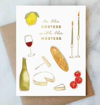 White greeting card featuring watercolor artwork of a wine glass and bottle, corkscrew, cheese wheel and wedge, baguette, tomatoes, taper candles, and a lemon says, "To the hostess with the mostess" in gold foil at top center; a kraft paper envelope rests underneath