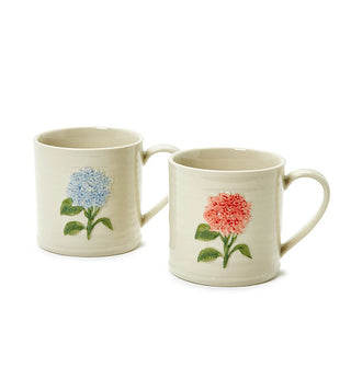 Two cream-colored stoneware mugs, one with a blue hydrangea flower, and the other with a reddish-pink hydrangea flower, both in a hand-painted embossed style with green stems and leaves