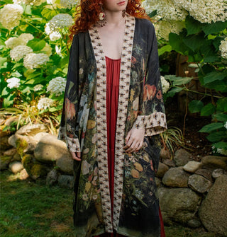 Model standing in a garden wears a long, kimono-style robe with floral motif and contrasting bee print borders