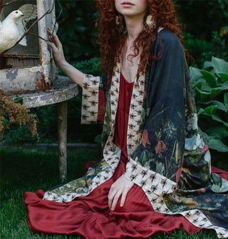 Model sitting on the grass in a garden wears a long, kimono-style robe with floral motif and contrasting bee print borders over a red dress