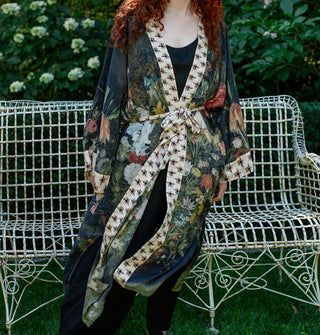 Model standing in front of an antiqued white wrought-iron bench wears a long, kimono-style robe with floral motif and contrasting bee print borders, tied at the waist with a matching fabric belt