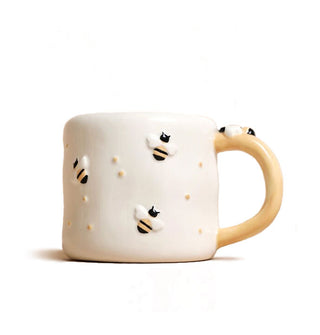 Slightly irregularly shaped white ceramic coffee mug with peachy-colored handle features raised and painted bumblebee details and small gold bumps across its surface