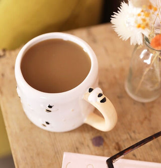 White coffee mug with raised bumblebee details is filled with a brown beverage and sits on a rustic wooden tabletop next to a small clear glass vase of white and orange flowers