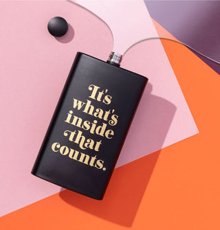 Black flask with gold lettering that says, "It's what's inside that counts" rests on a pink, orange, and purple surface with its lid removed and a clear section of acrylic positioned next to it to suggest liquid pouring out