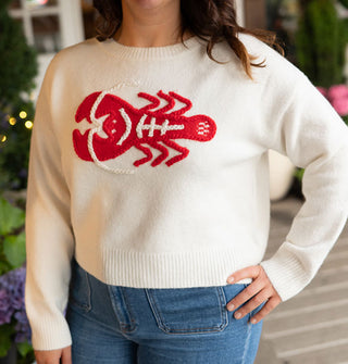Model with hand on hip wears a cream-colored long-sleeved sweater with a large red and white embroidered lobster design on the front