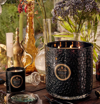 Lit multi-wick Jardin de Verveine candle in textural black glass vessel rests on a  wooden tabletop with another, smaller Voluspa candle, fresh flowers, glassware, and implements.