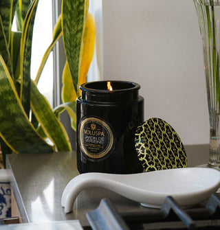 Lit Jardin de Verveine Voluspa candle in black glass jar with patterned tin lid removed and propped up next to it are staged next to a gas range with Dracaena trifasciata in the background