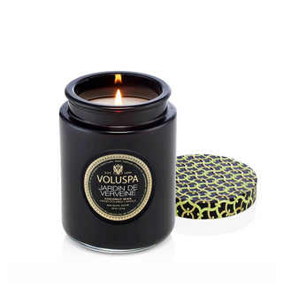 Lit single-wick Jardin de Verveine Voluspa candle in black glass vessel with patterned tin lid removed and set to the side