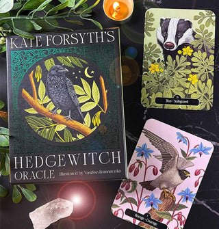 Kate Forsyth's Hedgewitch Oracle card deck box and two sample cards (Rue: Safeguard and Borage: Courage) staged with plants, lit candle, and quartz stone