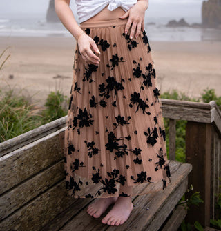 Model wears a pleated tan tulle skirt with black floral embroidery on a beach boardwalk