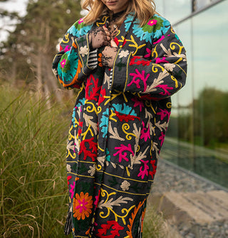 Model wears a colorfully embroidered black jacket with striped lining in front of a glass structure in a wooded, grassy setting