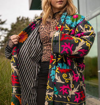 Model wears a colorfully embroidered black jacket with striped lining in front of a glass structure in a wooded, grassy setting
