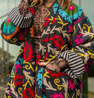Model wears a colorfully embroidered black jacket with striped lining in front of a glass structure