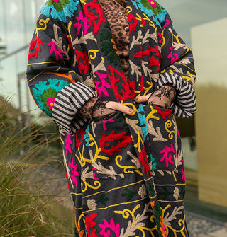 Model places lace-gloved hands in the pockets of a colorfully embroidered black jacket with striped lining in front of a glass structure in a wooded, grassy setting