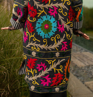 Model wears a colorfully embroidered black jacket with striped lining in front of a glass structure in a grassy setting
