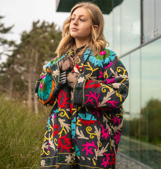 Model wears a colorfully embroidered black jacket with striped lining in front of a glass structure in a wooded, grassy setting