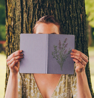Model sitting in front of a tree holds an opened lavender embroidery journal in front of face
