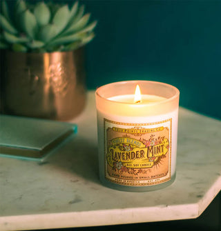 A lit Lavender Mint candle with decorative label rests on a marble tabletop with a book and succulent in the background