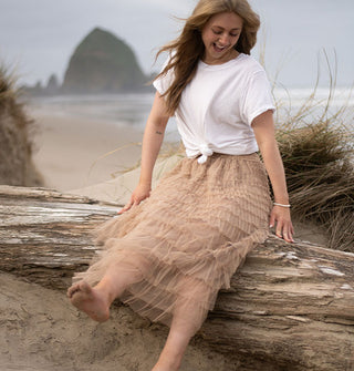 Smiling model sitting on driftwood wears a long beige tiered tulle skirt with knotted white T-shirt against a beach backdrop