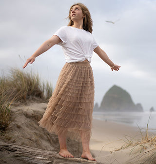 Model with arms outstretched wears a long beige tiered tulle skirt with knotted white T-shirt against a beach backdrop