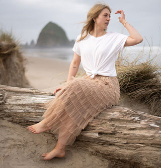 Model sitting on driftwood with arm raised wears a long beige tiered tulle skirt with knotted white T-shirt against a beach backdrop