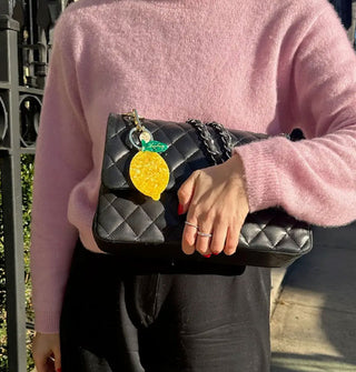 Model in pink sweater and black pants holds a quilted black purse with lemon keychain attached