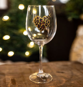 Clear wine glass with a rhinestone-encrusted leopard print heart decal sits on a wooden surface against a lit tree backdrop