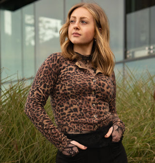Model with hands in the pockets of black pants wears a leopard print button-down cardigan in front of tall grass and a glass structure