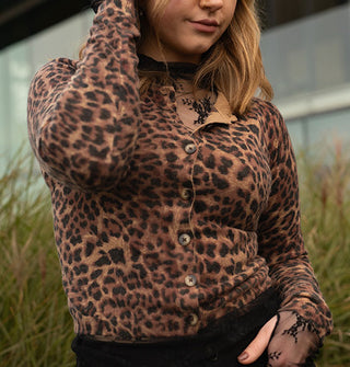 Model wears a leopard print button-down cardigan with black sheer lace top underneath in front of tall grass and a glass structure