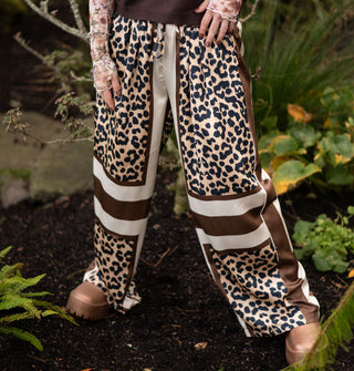 Model standing in a forest setting wears satin pants with leopard print blocks separated by brown and cream vertical and horizontal stripes