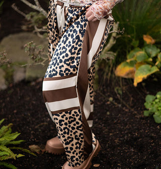 Model standing in a forest setting wears satin pants with leopard print blocks separated by brown and cream vertical and horizontal stripes