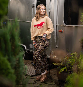 Smiling model posing in front of an Airstream trailer wears leopard print pants paired with a tan sweater with red galloping horse design and copper clogs