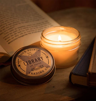 Lit Library jar candle is staged two closed and one opened book on a wooden surface