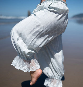 Model wearing nearly white, light blue, lace-trimmed pantaloons lifts one leg, bent at the knee