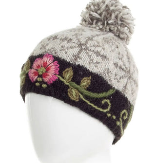 White beanie with pink and green floral embroidery on black around the band, beigey-gray snowflake pattern around the crown, and a gray and white pom pom at the top