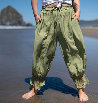 Model standing on the beach with sea stacks behind wears a pair of green, lace-trimmed pantaloons with a knotted white T-shirt