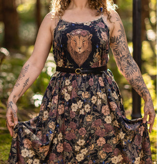 Model holds the skirt of a sleeveless floral patterned dress featuring a lion's head on the bodice and styled with a thin black belt with gold buckle