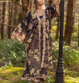 Model standing in a mossy forest next to a lamp post wears a floral print robe with contrasting  borders