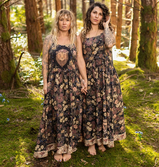 Two models standing in the woods and holding hands each wear a floral patterned full-length sleeveless dress, one with a lion's head on the bodice