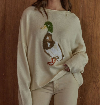 Model standing against a woodgrain backdrop wears a cream-colored sweater with mallard graphic
