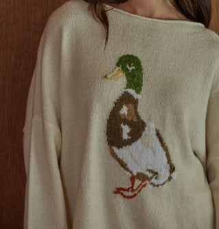 Model standing against a woodgrain backdrop wears a cream-colored sweater with mallard graphic