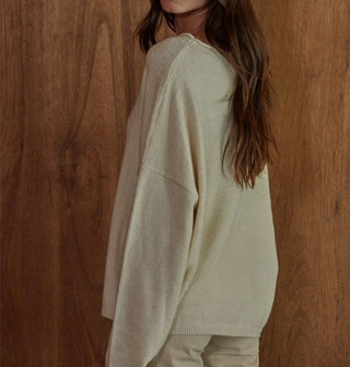 Model standing in front of a woodgrain backdrop and turning away from the camera wears a cream-colored sweater