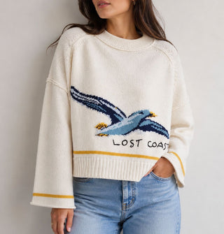 Model wears a cream-colored sweater with wide neckline, drop shoulder, and bell sleeves that features a large seagull graphic and the words, "Lost coast" near the bottom, accented by a yellow stripe above the bottom ribbed hem and at the rolled sleeve openings