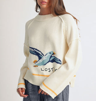 Model wears a cream-colored sweater with wide neckline, drop shoulder, and bell sleeves that features a large seagull graphic and the words, "Lost coast" near the bottom, accented by a yellow stripe above the bottom ribbed hem and at the rolled sleeve openings