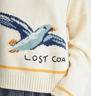 Closeup of the blue and yellow seagull graphic, "Lost coast" lettering, and yellow stripe on a white sweater with ribbed bottom hem