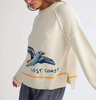 Model wears a cream-colored sweater with wide neckline, drop shoulder, side slit, and bell sleeves that features a large seagull graphic and the words, "Lost coast" near the bottom, accented by a yellow stripe above the bottom ribbed hem and at the rolled sleeve openings